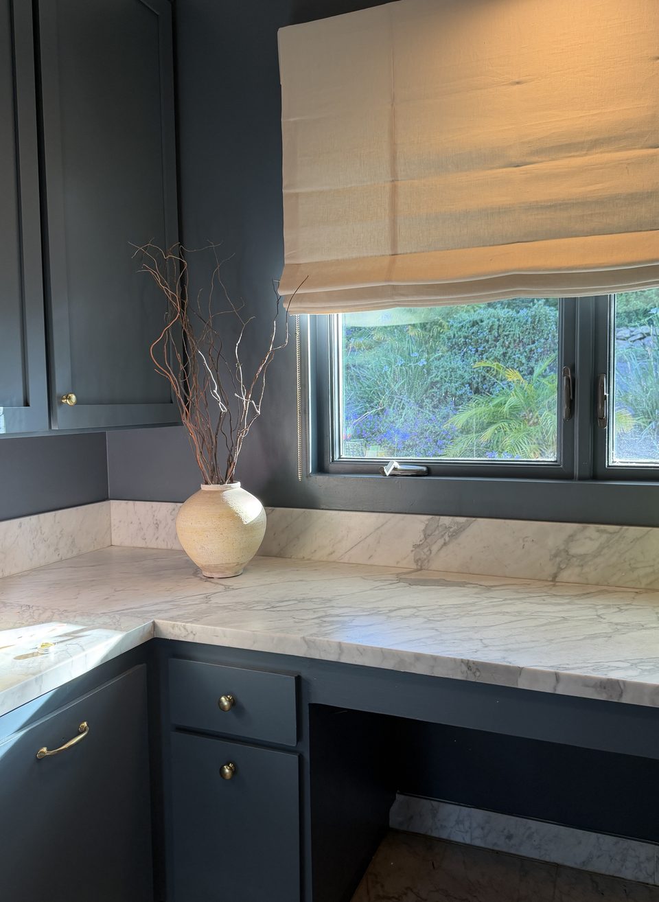 Dark cabinetry with white marble countertop, ceramic vase, and garden light through a window